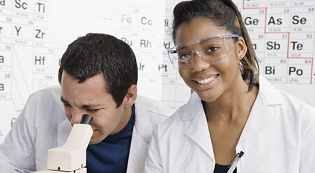 two students looking into a microscope with periodic table in background