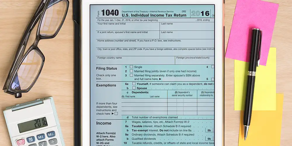 A tablet displaying form 1040, with glasses, a calculator, pen and post it notes on the sides of it.