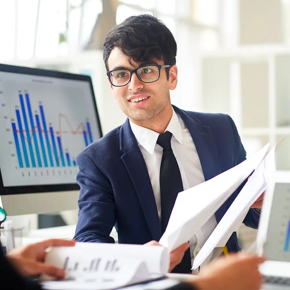 Economist holding papers near a computer, discussing with another person in the foreground.
