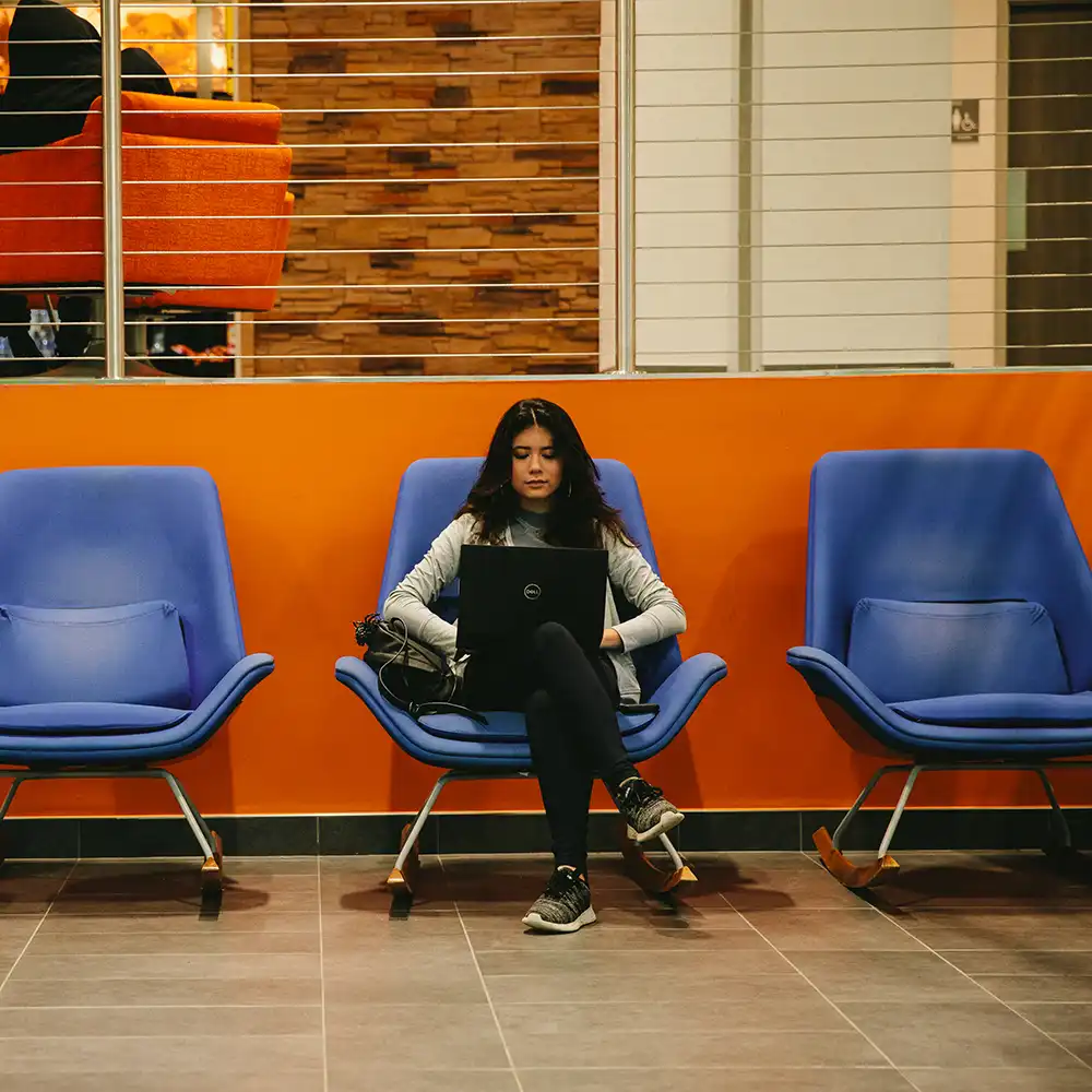 A student working on their laptop on a blue chair.