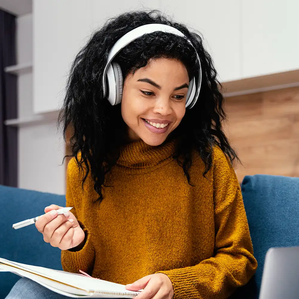 Student looking at their laptop with headphones, about to write in a notebook.