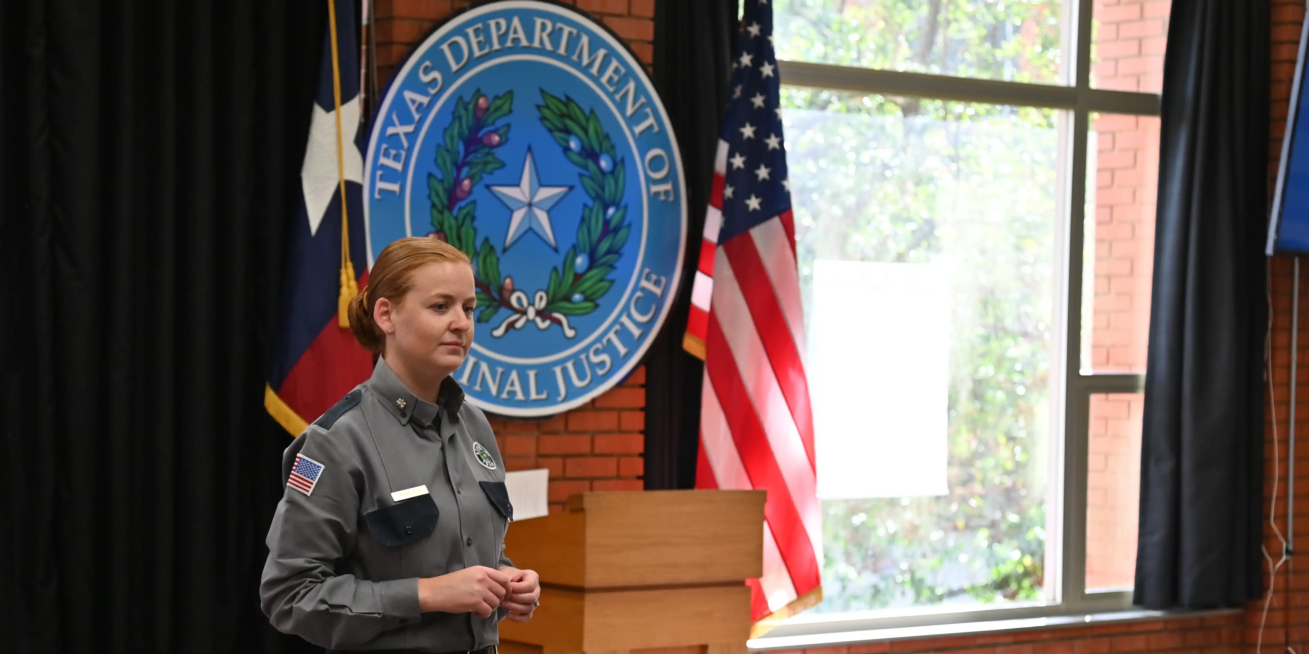 Corrections officer in uniform in training room listening to participant (off camera). The Texas Department of Criminal Justice seal can be seen in the background.