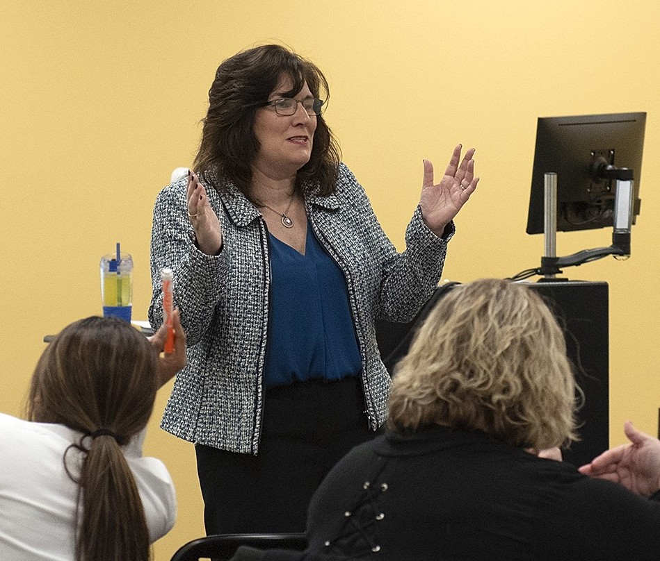 Professor speaking to the camera in a classroom style class, with her hands opened.