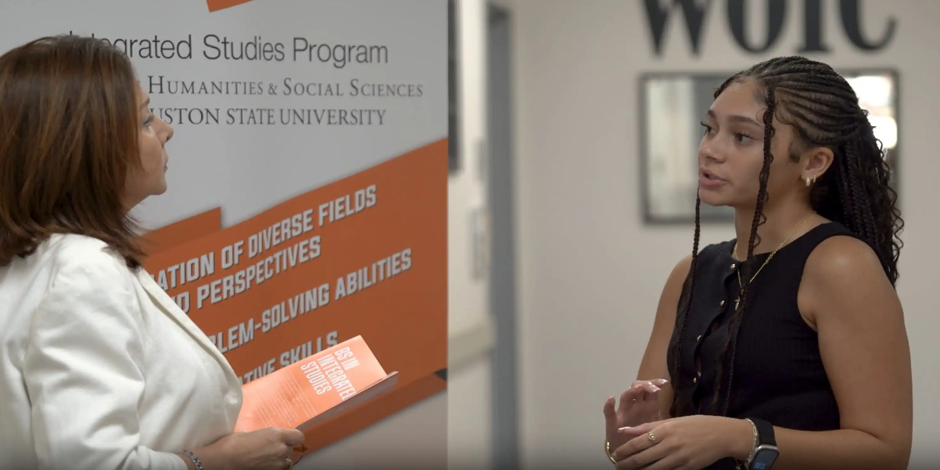 SHSU student speaks with faculty member in front of a banner for the Integrated Studies program.
