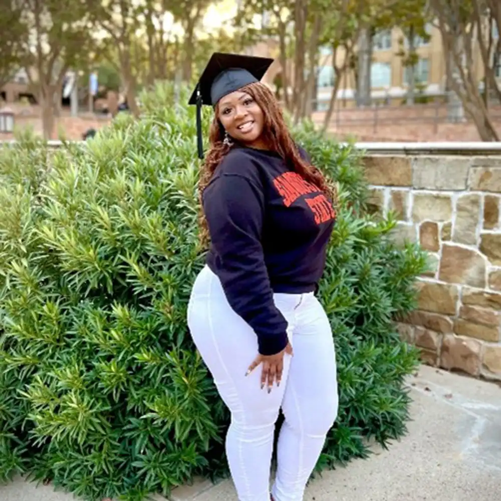 Trichell Spikes poses outside in her commencement hat, SHSU sweater, and white pants on campus.
