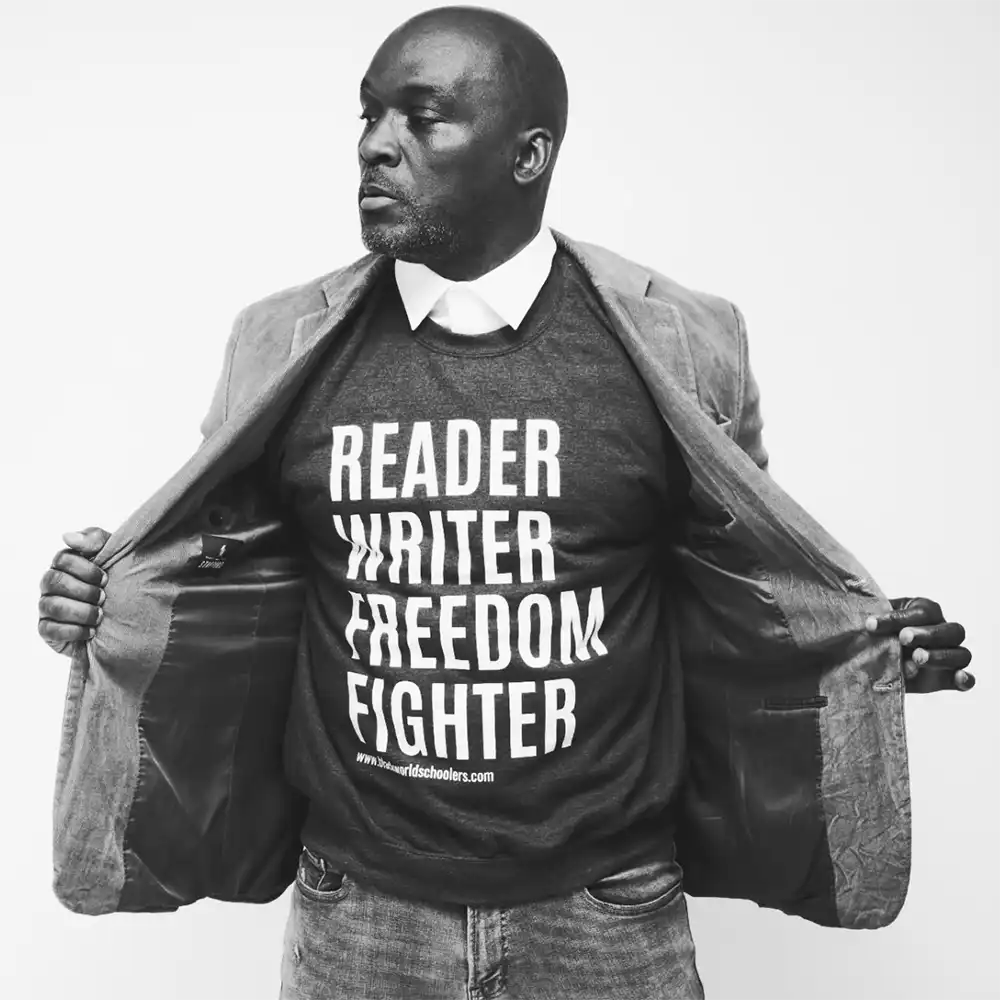 Derrick stand defiantly looking away from the camera holding his jacket open, his shirt reads "reader, writer, freedom fighter". 