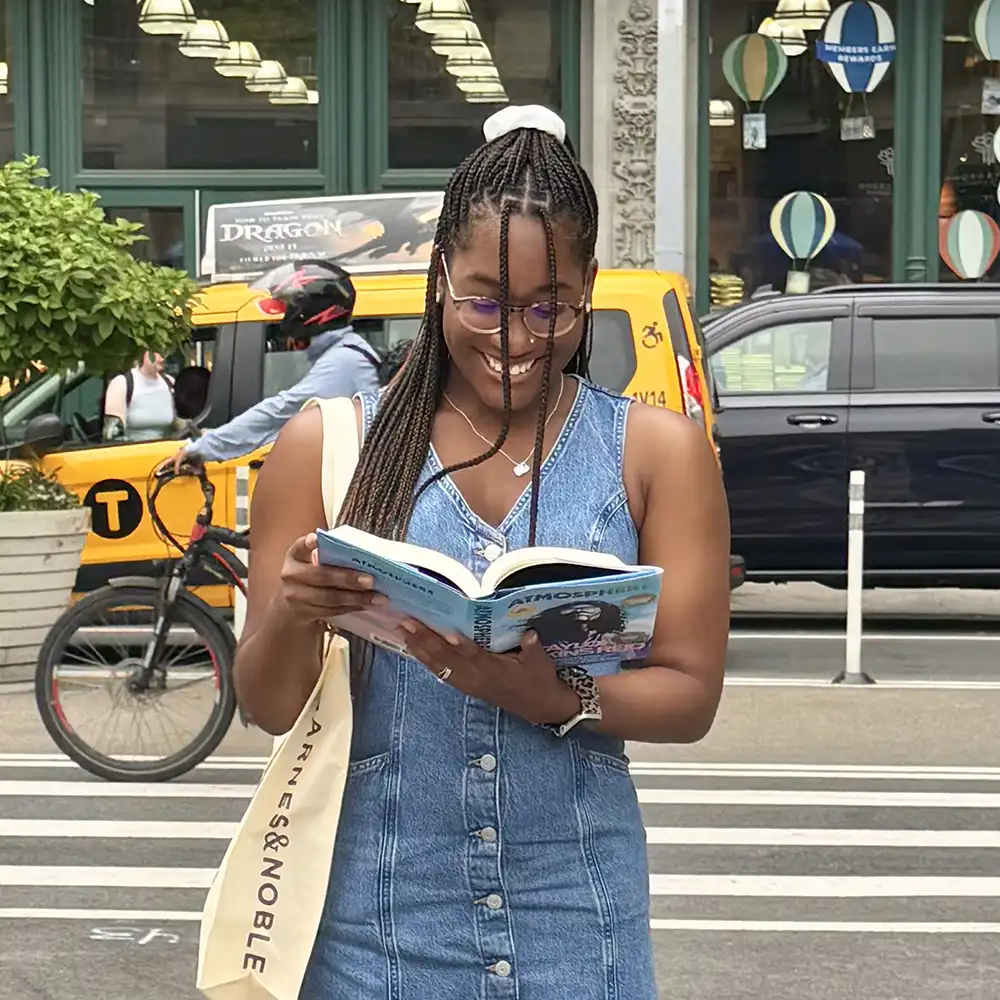 Lakyndra stands on a busy New York City street looking down at an open book with a wide smile.