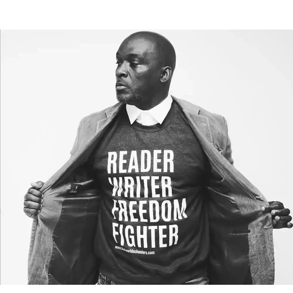 Derrick stand defiantly looking away from the camera holding his jacket open, his shirt reads "reader, writer, freedom fighter". 