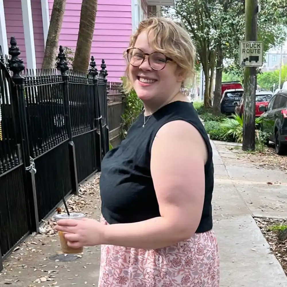 Bleah stands on a sidewalk in a New York City neighborhood smiling with an iced coffee in her hand.
