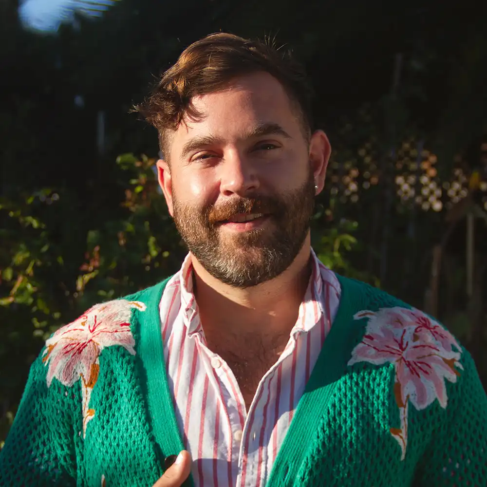 Sam smiles at the camera surrounded by greenery and in a bright green floral sweater.