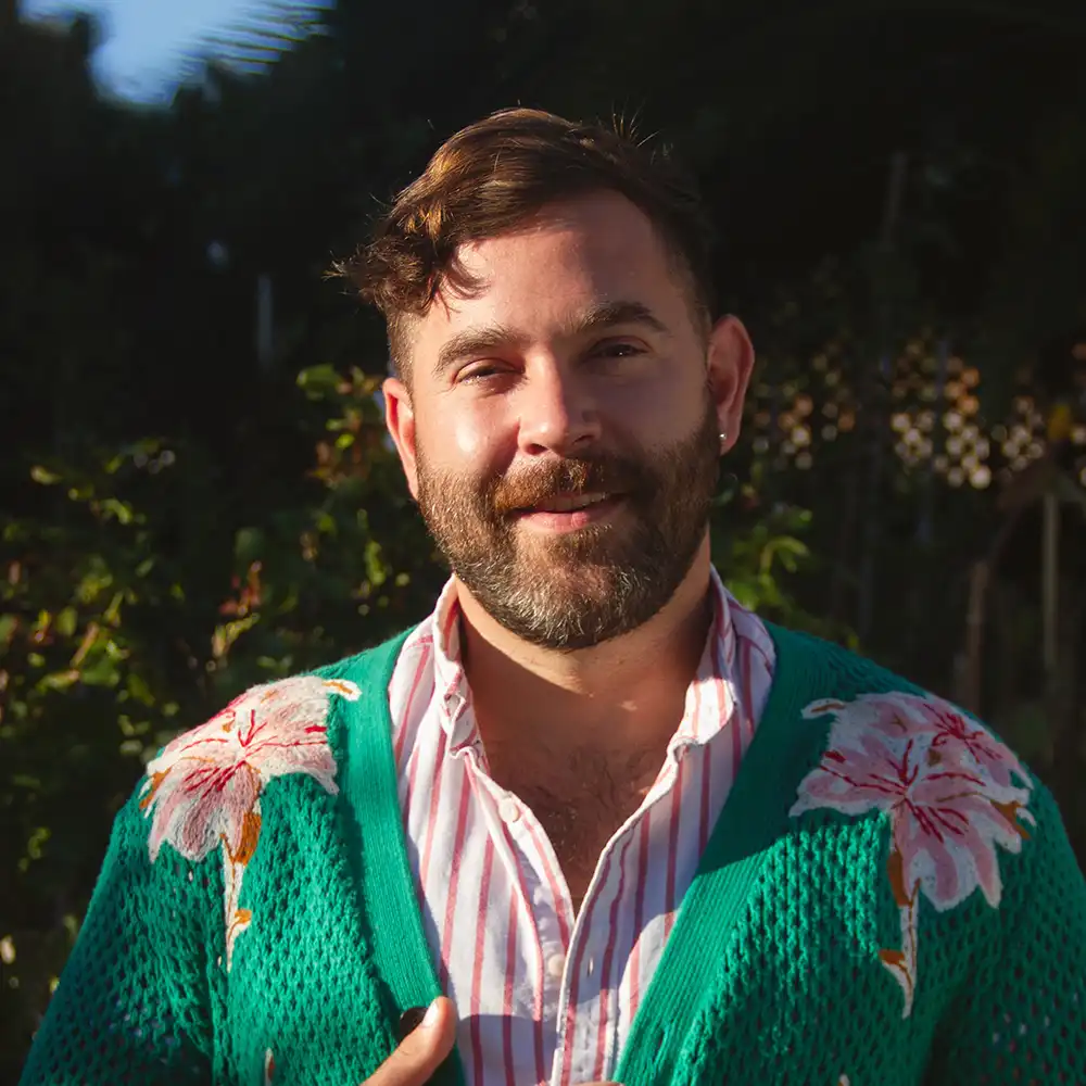 Sam smiles at the camera surrounded by greenery and in a bright green floral sweater.