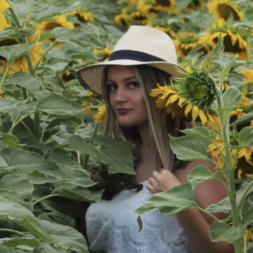 Olivia stands in a sunflower field with a sunhat on, looking at the camera.