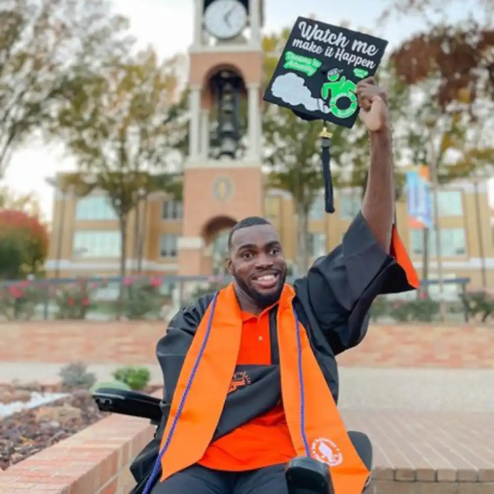 Broderick smiles in his graduation robes and holds up his motorboard which reads 'watch me make it happen'.