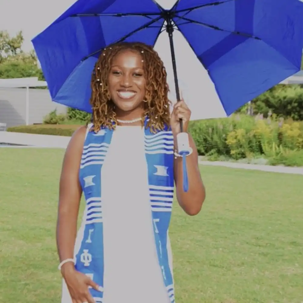 A'Shari smiles and poses on campus in her graduation outfit holding a blue and white umbrella.
