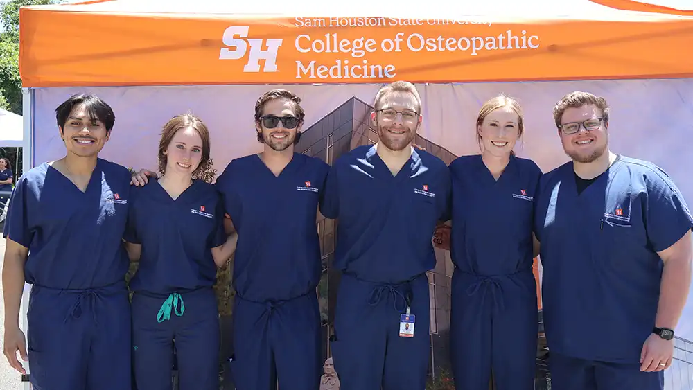 A group of SHSU College of Osteopathic Medicine student doctors in navy scrubs smile together in front of an orange SHSU-COM tent at an outdoor event.