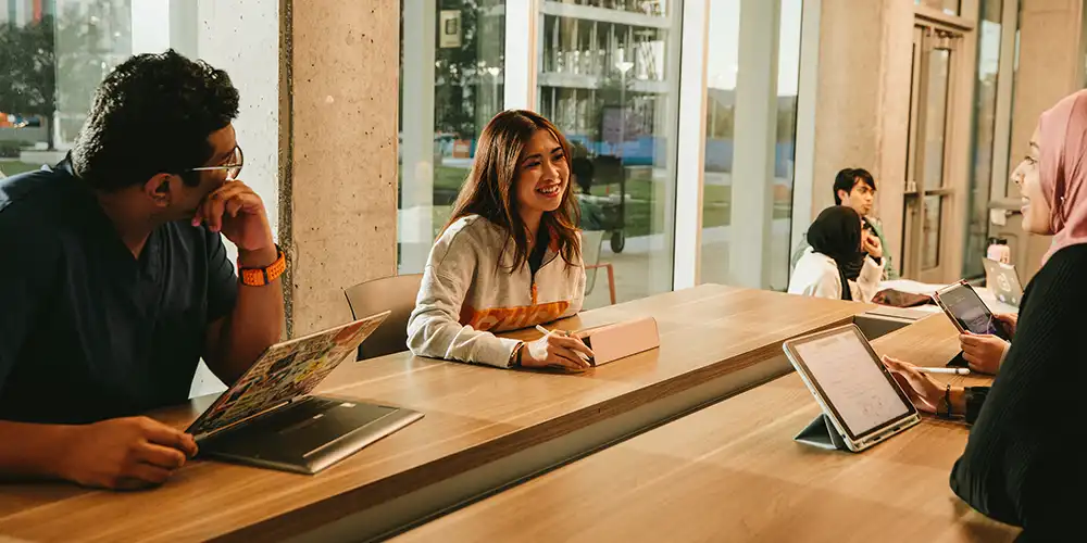 Student doctors sitting at a table and talking inside the COM building with computers and iPads in front of them