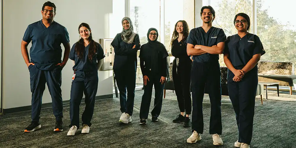 Group of seven students standing next to each other in a line in scrubs in front of windows