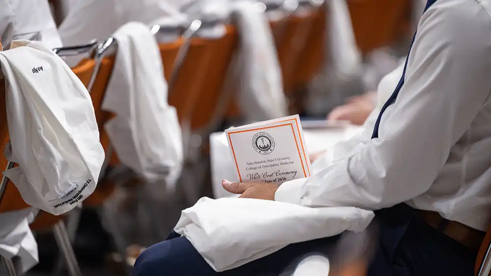 Detail shot of hands holding a white coat program with a white coat draped over a chair