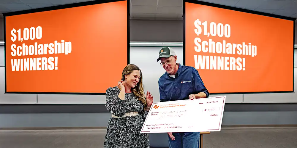 A student receives a $1,000 scholarship check during an orientation raffle event, standing with a presenter in front of large orange screens that read “$1,000 Scholarship Winners!”