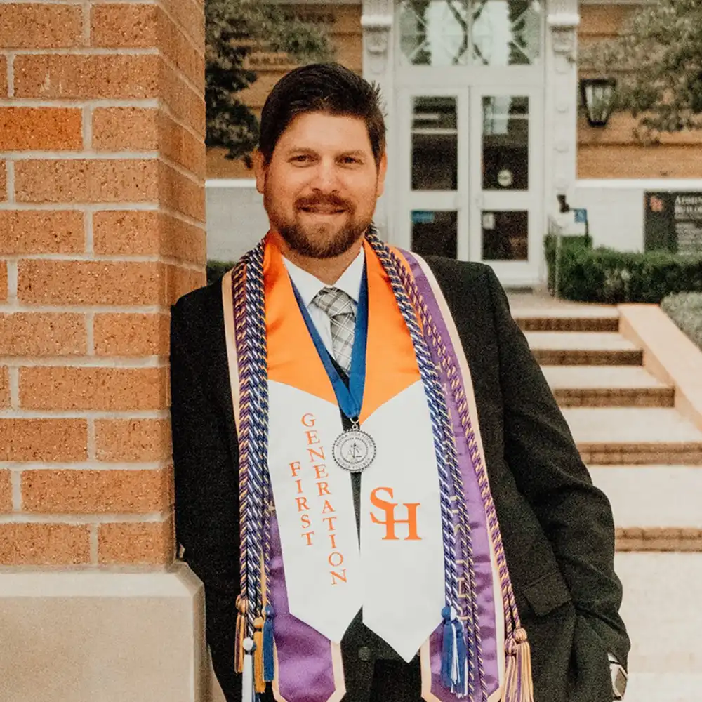 SHSU student leaning their should on the brick of the clock tower on campus wearing their graduation robes with many awards.