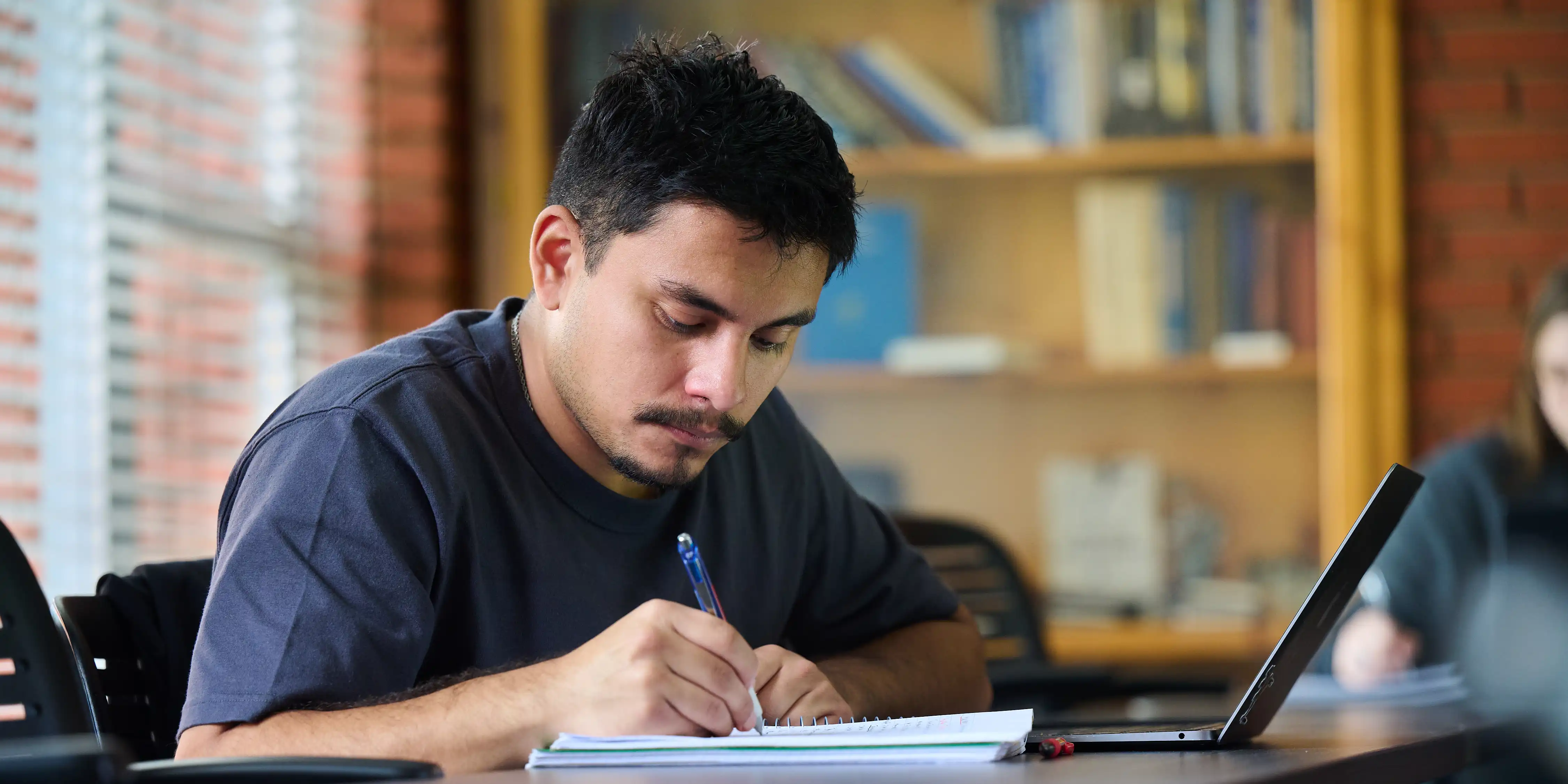 Student in classroom taking notes in a notebook with a laptop nearby.