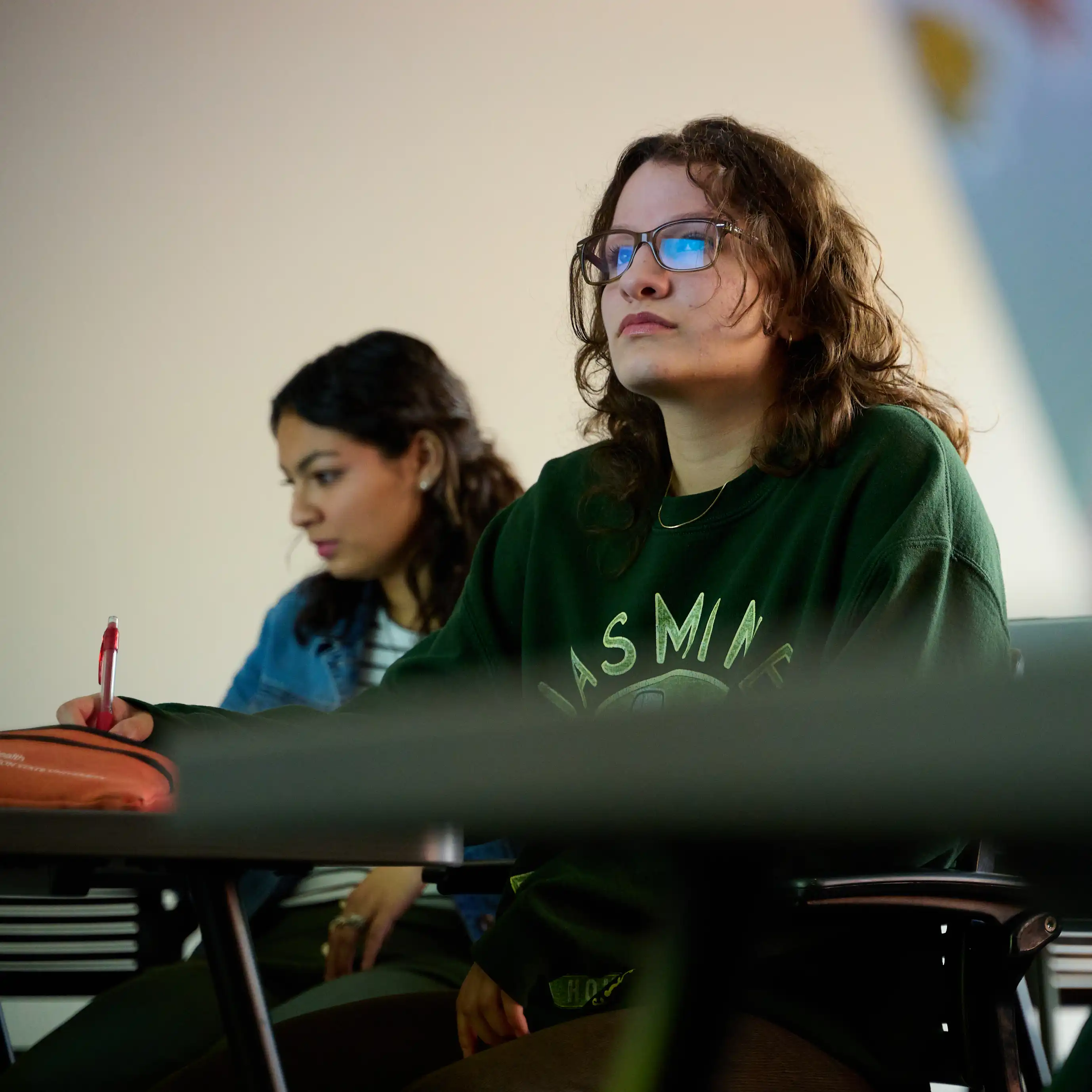 Two students in classroom setting listening to lecture and taking notes.