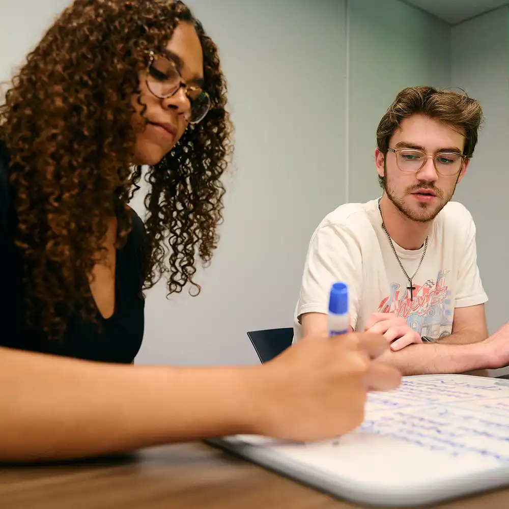 Undergraduate students writing on whiteboard on table in classroom.