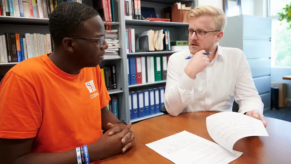 Professor speaking to the camera in a classroom style class, with her hands opened.