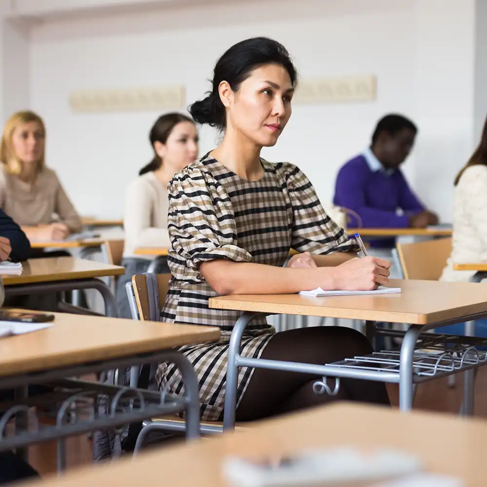 ​​Picture of adult students sitting in a classroom, in their own desks. Picture is focused on a lady, and all other students are looking away from the picture, to what seems the front of the class.​ 