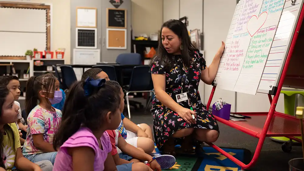 Teacher on her knees, by a whiteboard, taking to her students who are around her sitting down