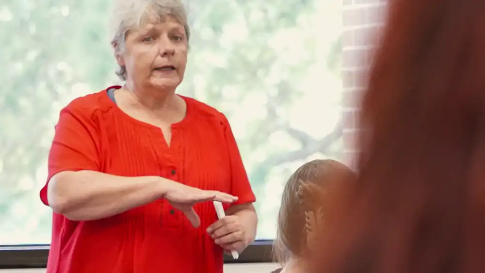 Professor speaking to the camera in a classroom style class, with her hands opened.