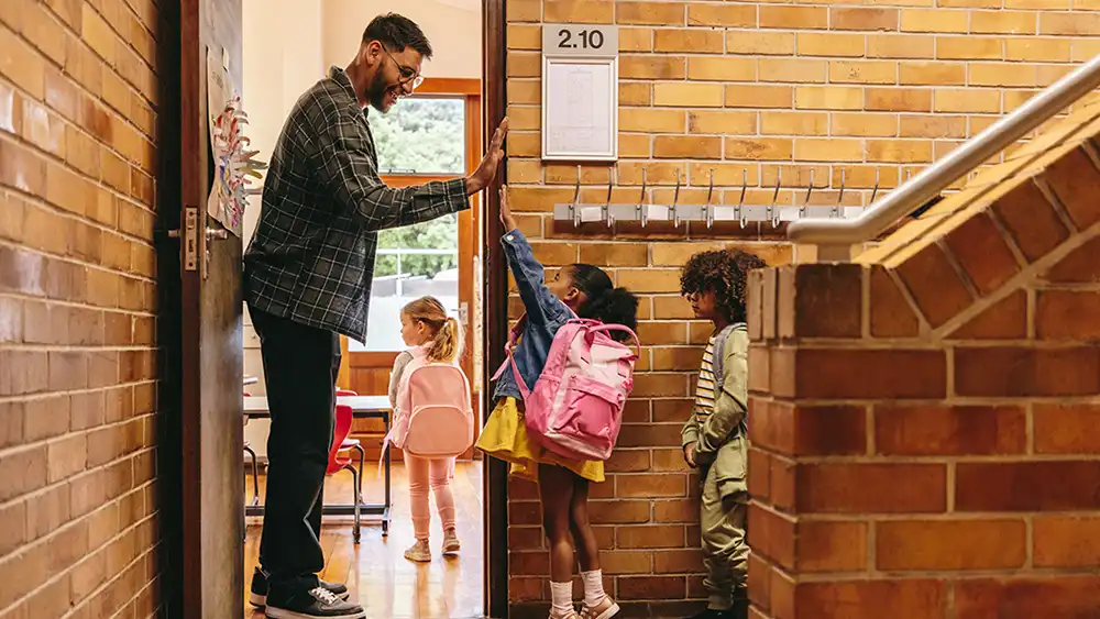 Male teacher greeting young students in the classroom hallway.
