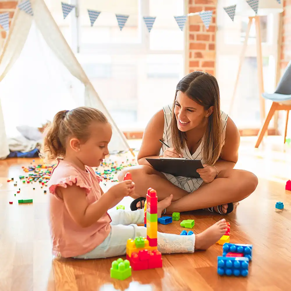 A lady is sitting down, smiling towards a little girl, leaning towards her, writing down on a notepad, while the little girl is sitting down, playing with Legos.