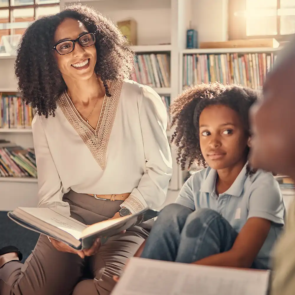 Woman with glasses reading with a book open, and 3 kids around her, all looking at one of the kids. 