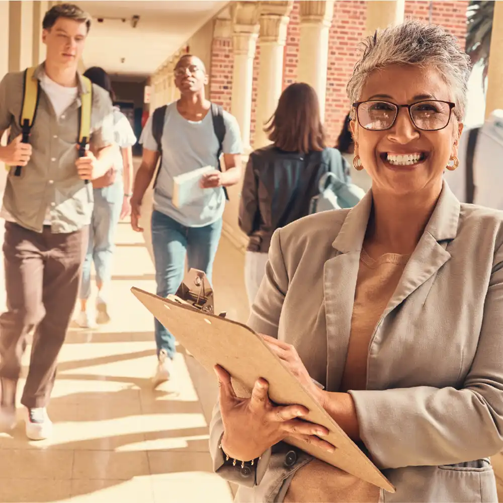 Lady holding a clipboard standing in a covered outdoor walkway and students are walking past.