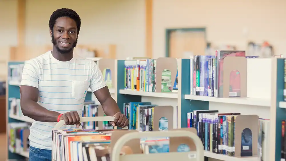 Man pushing cart of books in a library, smiling.