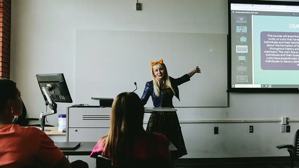 Professor speaking to the camera in a classroom style class, with her hands opened.