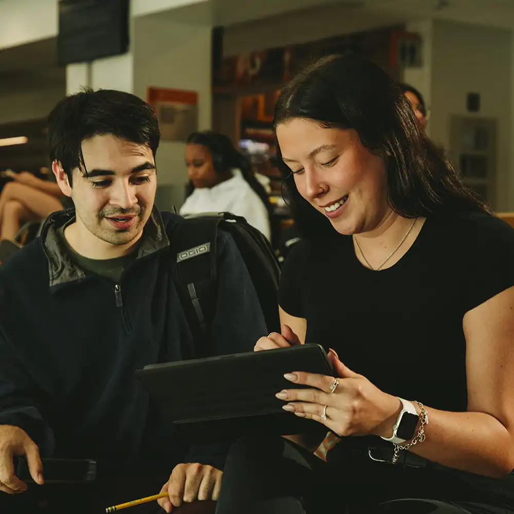 Two women looking at each other, smiling, sitting down, working on an assignment.