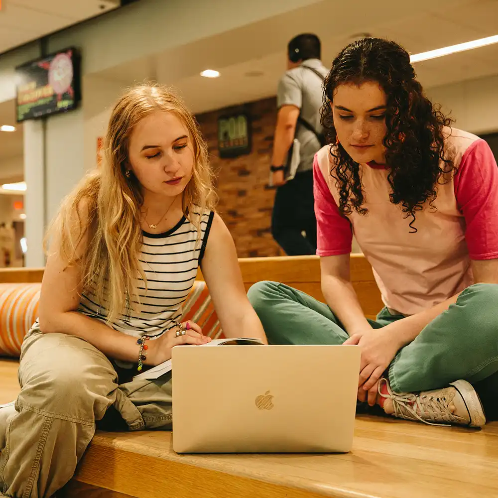 Two women looking at each other, smiling, sitting down, working on an assignment.