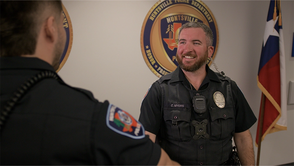 Male police officer shaking hands with his colleague, wearing his police officer uniform and standing in the station in front of a city seal on the wall