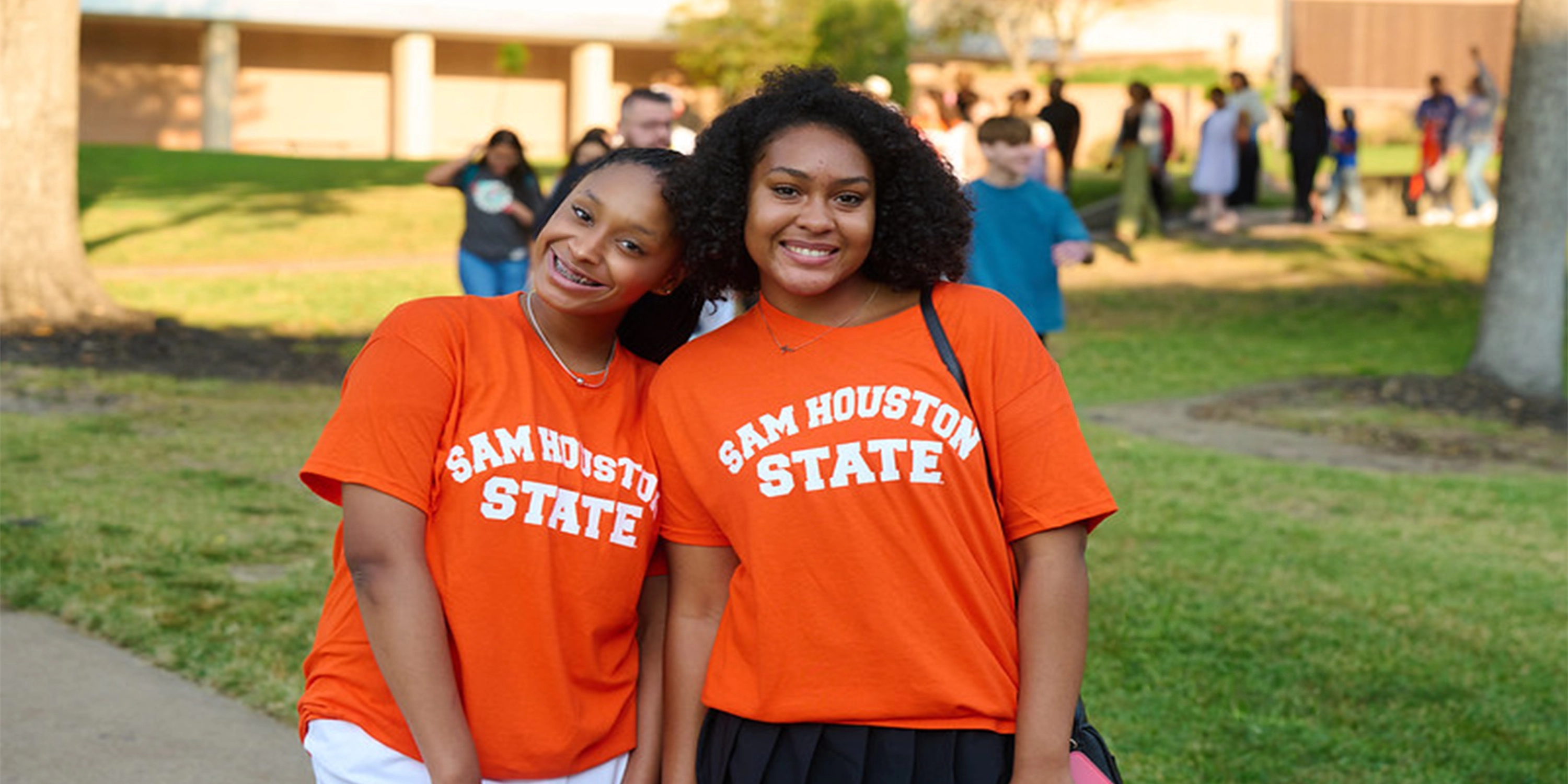 Two smiling Sam Houston State students standing outdoors on campus wearing orange SHSU shirts and posing together during a student event.