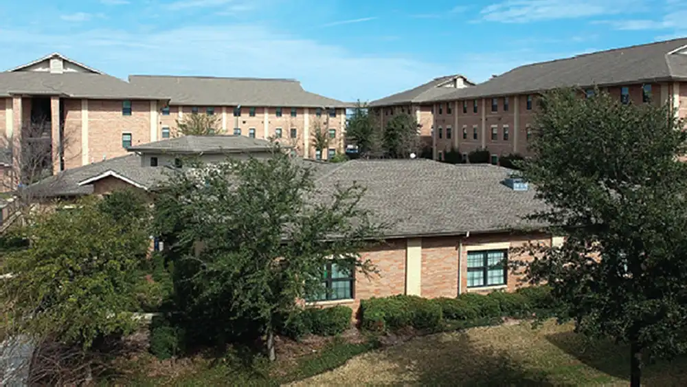 Exterior view of Bearkat Village Apartments at Sam Houston State University. In the foreground is a single-story clubhouse with a grey roof and large windows, surrounded by neatly trimmed bushes and trees. Behind the clubhouse, three multi-story residential buildings with light brown brick exteriors and grey roofs are visible. The sky is clear and blue with a few wispy clouds.