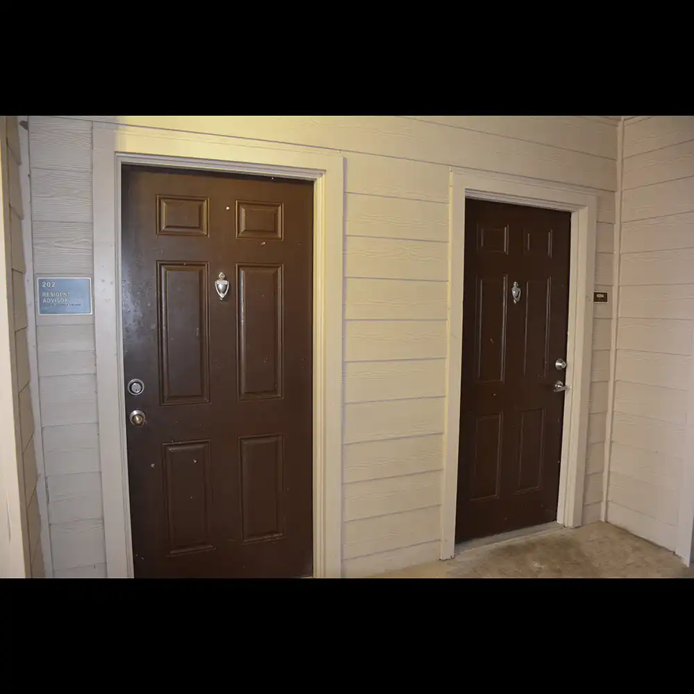 The image shows the exterior of two apartment doors at SHSU's Bearkat Village Apartments. Both doors are dark brown with silver door handles and peepholes. The door on the left has a small blue sign next to it that reads "202 Bearkat Village Apartments." The walls surrounding the doors are beige with horizontal siding.