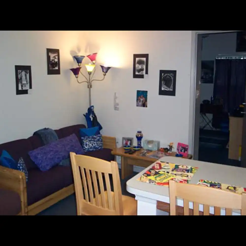 The living room at SHSU's Bearkat Village Apartments features a cozy setup with a dark maroon couch adorned with blue and purple pillows. Above the couch are three framed black-and-white photographs. A colorful floor lamp with multiple shades stands beside the couch, next to a small wooden side table holding a water bottle and decorative items. In front of the couch is a wooden dining table with two chairs, covered in magazines and reading materials. The room has white walls and an open doorway leading to another room.