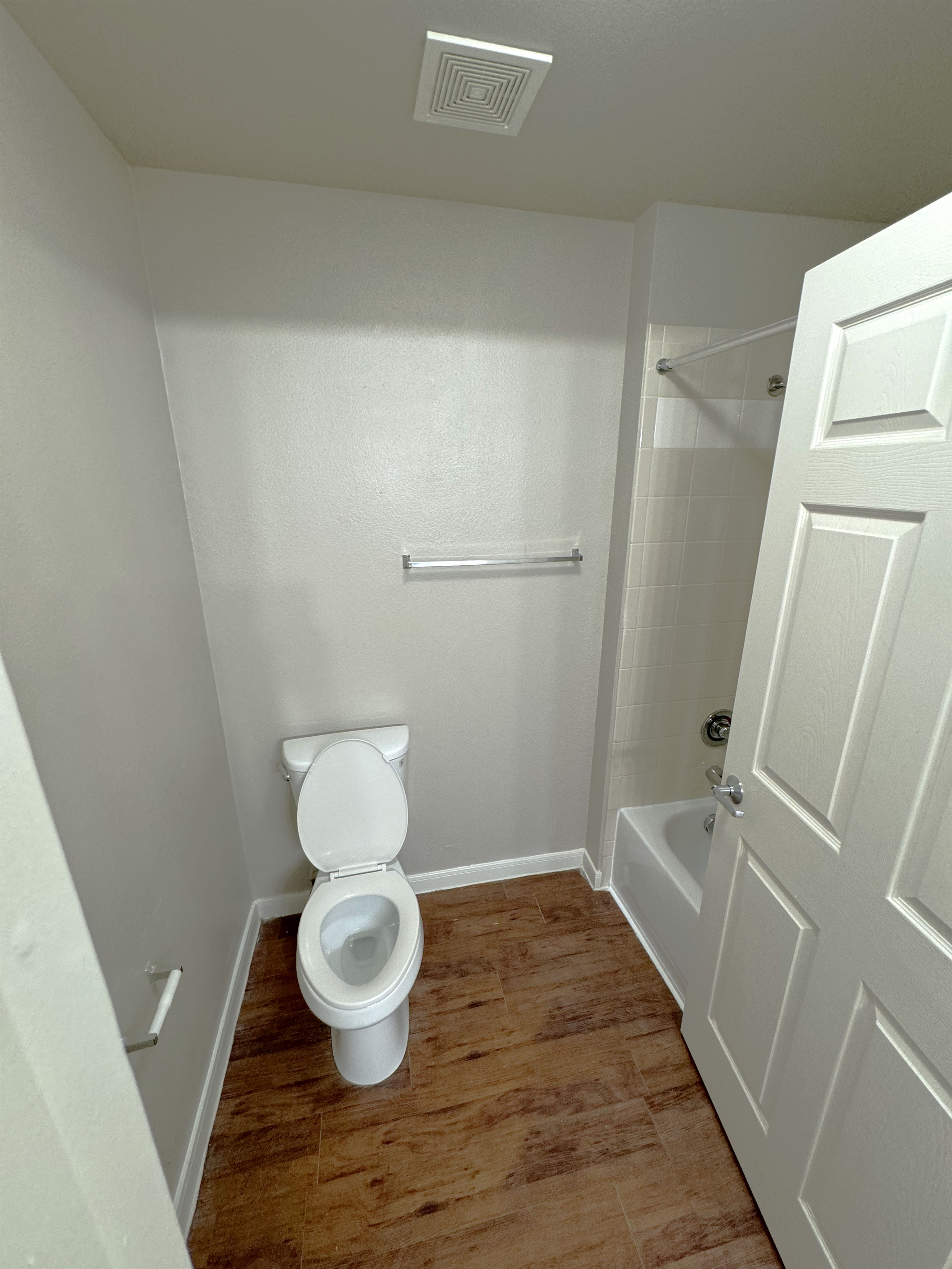 Bathroom at SHSU's Campus Edge Apartments featuring a white toilet with an empty towel rack above it, a bathtub with a shower head and white tiled walls, and wood-like vinyl flooring. The walls are painted light gray, and a ceiling vent is located above the toilet. An open door is visible on the right side of the image.
