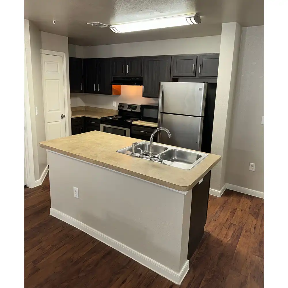 Modern kitchen at SHSU's Campus Edge Apartments featuring dark wooden cabinets, full-size refrigerator, electric stove with oven, and microwave. A large island with a beige countertop and double sink with chrome faucet sits in the center. The floor is dark laminate, walls are light gray, and a bright overhead light fixture illuminates the space.