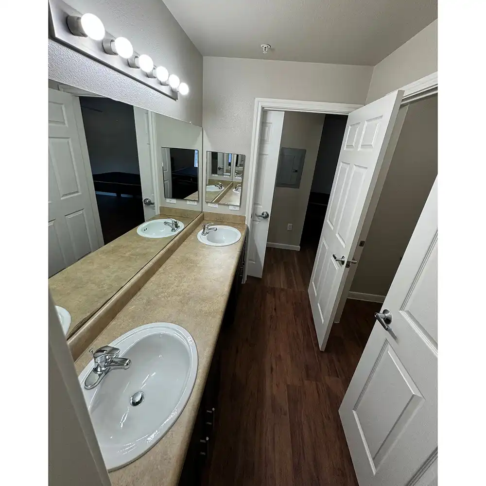 Jack-and-Jill vanity area at SHSU's Campus Edge Apartments featuring a long countertop with two white sinks and chrome faucets. A large mirror spans the length of the counter, with several round vanity lights mounted above it. The floor is covered in wood-like laminate. Two white doors are visible, one partially open and another reflected in the mirror.