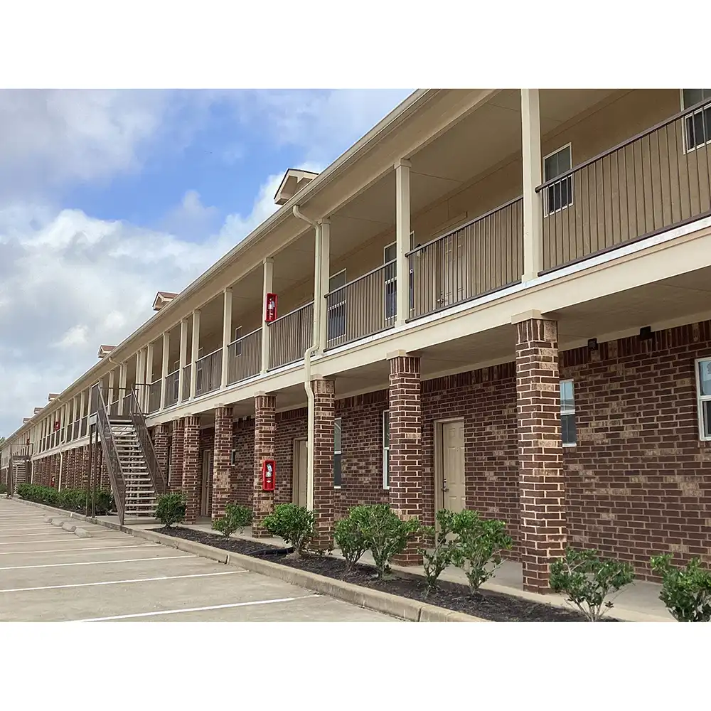 The image shows the exterior of one of the buildings at SHSU's Copper Village Apartments. The building is a two-story structure with a brick facade on the ground floor and beige siding on the upper floor. A long balcony runs along the upper level, supported by brick columns, with a metal staircase providing access. Small shrubs are planted in front, and a parking area with marked spaces is visible. The sky is partly cloudy.
