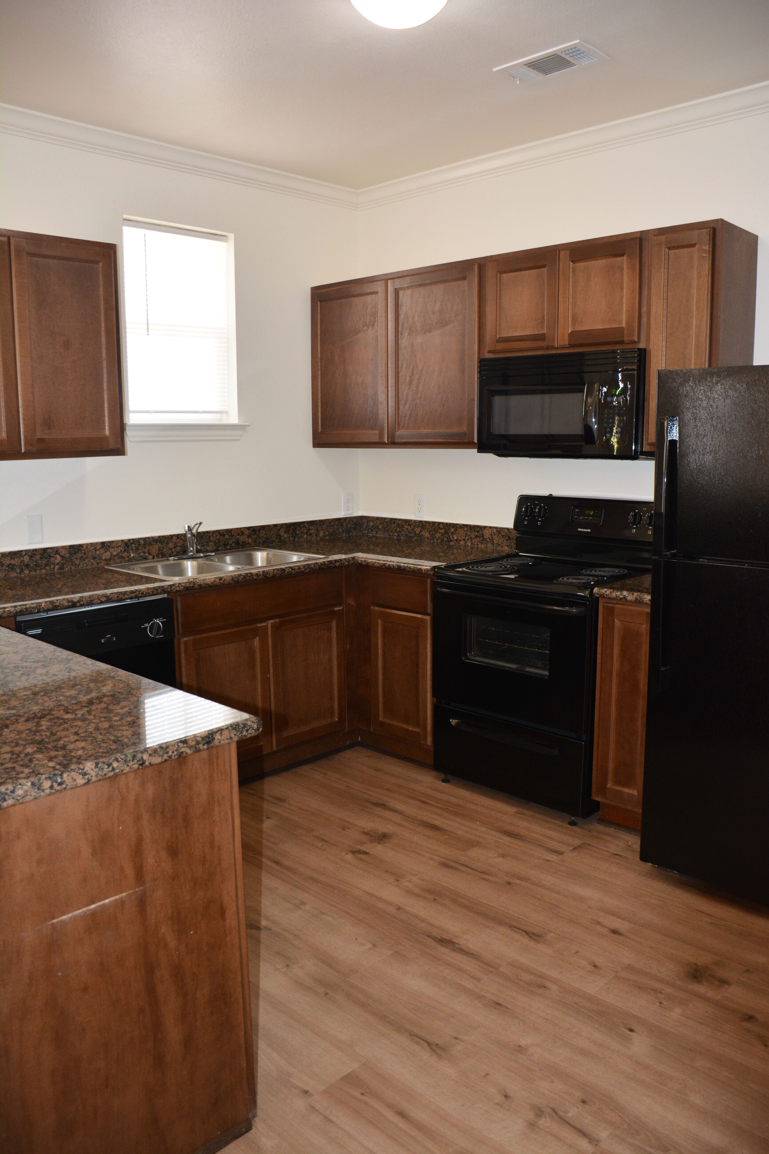Kitchen in SHSU’s Copper Village Apartments with wooden cabinets, granite countertops, and black appliances including refrigerator, stove, microwave, and dishwasher. A double sink sits beneath a small window, and the floor has a wood-look finish.