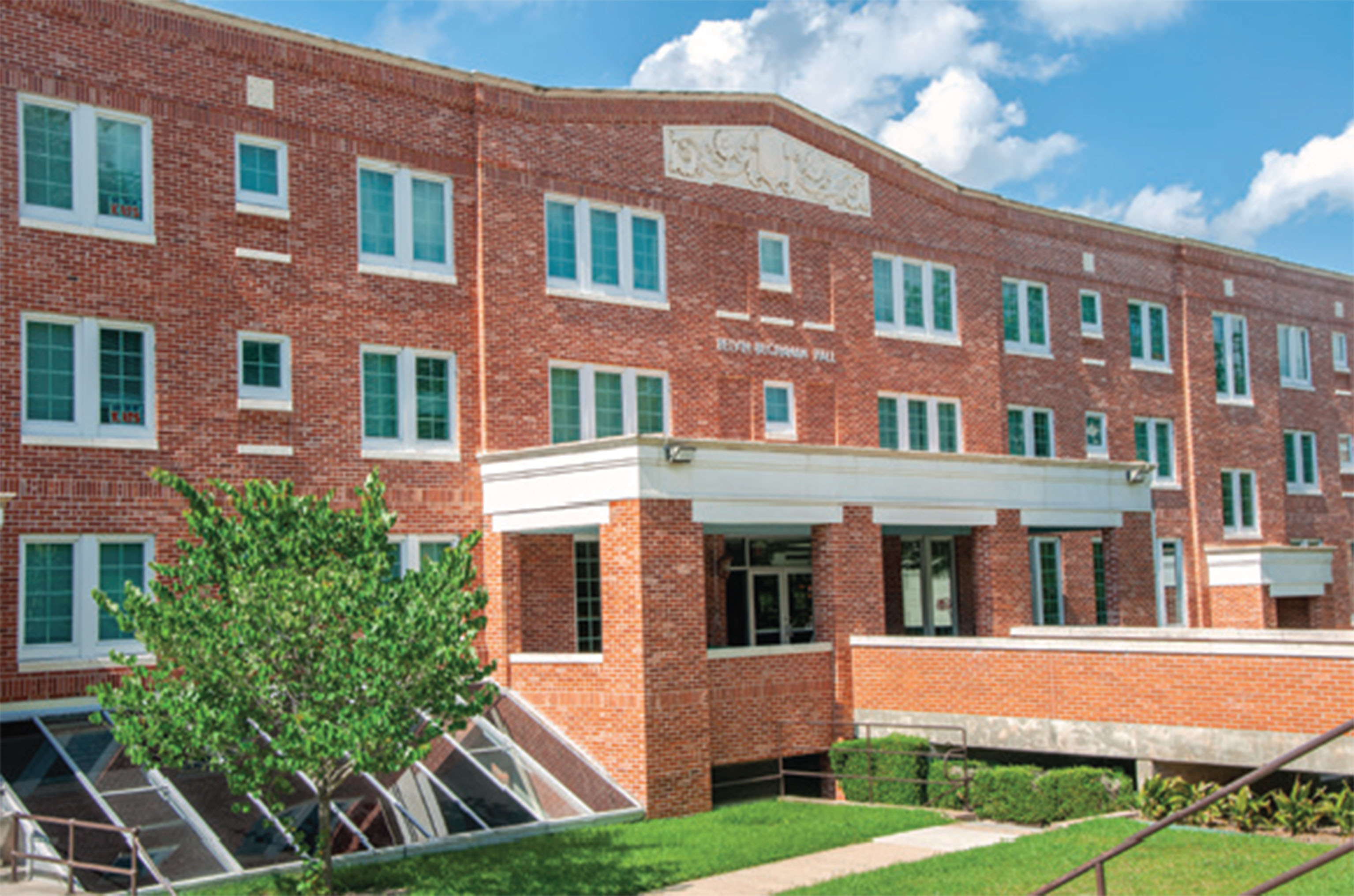 Exterior view of Belvin-Buchanan Hall, a red-brick residence hall with white-trimmed windows at Sam Houston State University, shown on a sunny day with blue skies and scattered clouds. A small tree and glass window structure are visible in the landscaped front lawn.